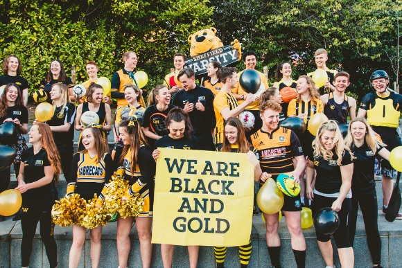 Members of sports teams stood in rows holding a banner saying We Are Black and Gold